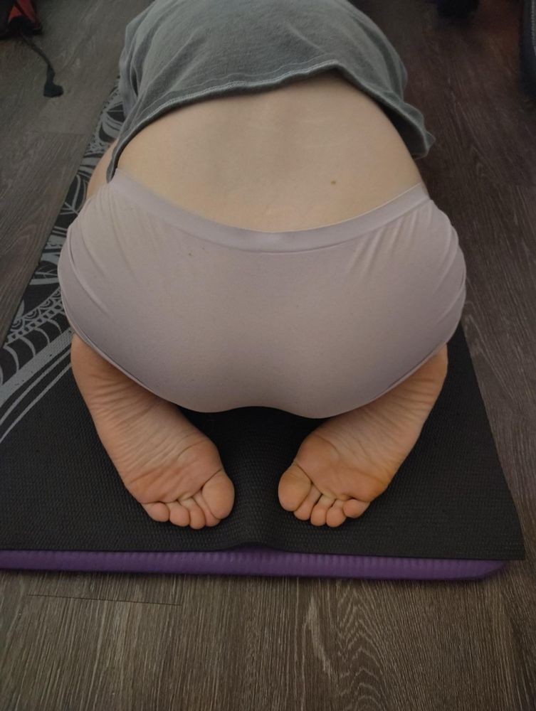 Luna in child’s pose shot from behind. Her legs are folded underneath her and her arms are clutching her sides as she rests and self-soothes. Her firm, tight ass is seated on the balls of her feet and her soles and toes are showing towards the camera