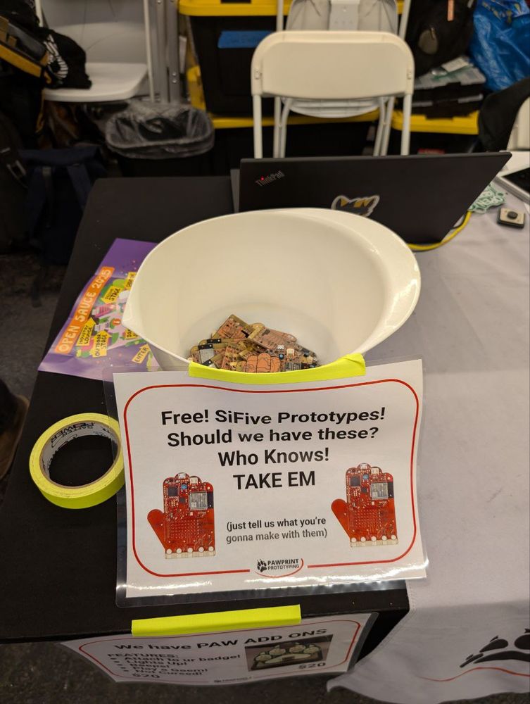 photo of a bowl on our exhibit table at open sauce 2025. bowl has orange hand shaped PCBs inside. the sign on the bowl has a photo of the hand shaped PCBs and reads: “ Free! SiFive Prototypes! Should we have these? Who knows! TAKE EM. (just tell us what you’re gonna make with them)” 