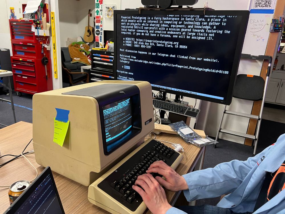 A user in a lab coat uses a VT100 to browse the web, the terminal displays the article for Pawprint Prototyping from the Noisebridge wiki.  The video output from the terminal is also displayed on a modern LCD TV.