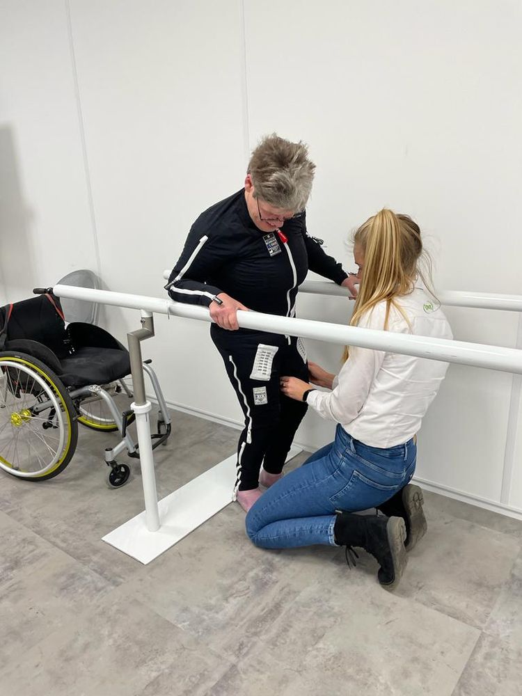 Vrouw in Exopulse Mollii suit staat met steun voor een geknielde jonge vrouw (fysiotherapeut). Een rolstoel staat achter haar. 