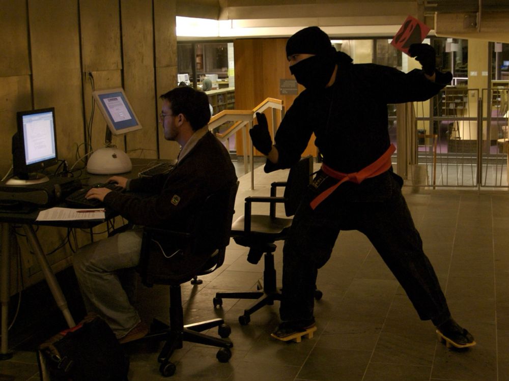 A young man sits typing at a computer in a college library. Behind him, a ninja wearing a black mask, gi, orange belt, and geta approaches, wielding a piece of construction paper with a ninja mask drawn on it.