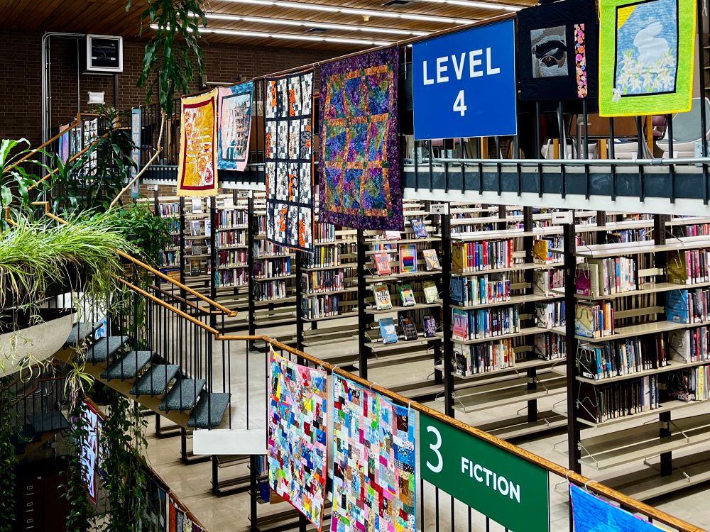 Quilts hanging from the railings in a library with book stacks and plants in the background