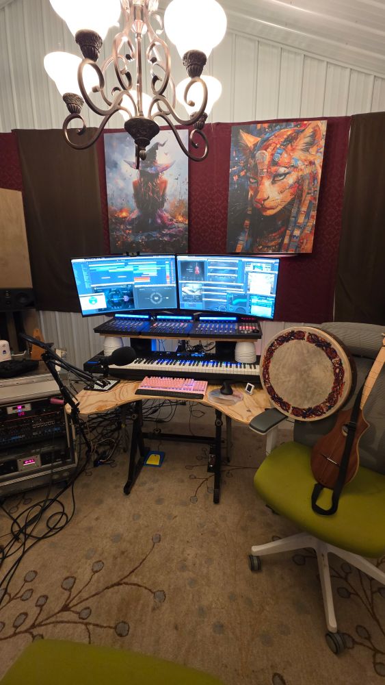 A wide view of my studio environment. In addition to the software and hardware pictured previously, this image shows a bank of hardware and outboard audio gear on the left, and a bohdran and stick dulcimer on the right. A chandelier illuminates the whole scene, and two paintings/acoustic panels keep watch over everything in the background.