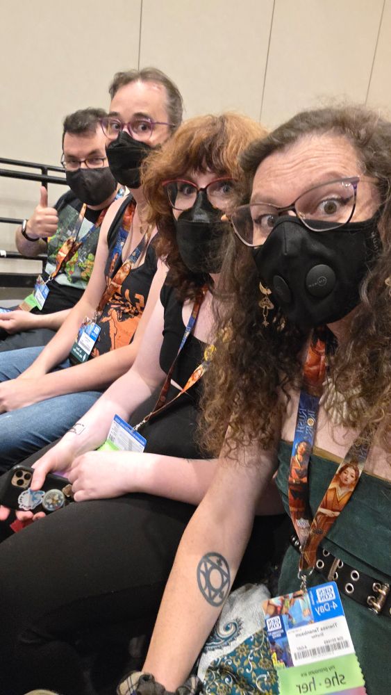 Three masked women, and one masked man sit next to each other in the audience for a panel. They all wear GenCon badges and looks of happy exhaustion on their faces.