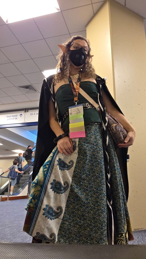A curly haired woman in a green patterned skirt, green bodice, and black cloak wearing a black mask, GenCon badge, and elf ears looks down at the camera, one hand on her thigh, the other resting on a paisley crossbody bag.