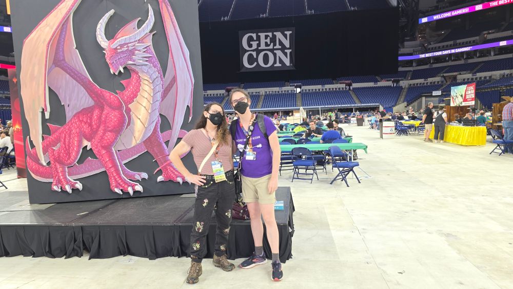 Two masked women stand in the Lucas Oil Stadium, arms around each other's waists, in front of a large banner with a red dragon on it. In the far background, a large GenCon banner is visible. Tables with gamers playing are arrayed behind them, and they are surrounded by the stadium bleachers. 