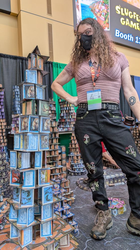A masked woman with curly hair stands next to a tower built out of Magic the Gathering cards that is almost as tall as she is. She wears black jeans with embroidered forest animals and mushrooms on them, and a mauve top, and Gencon badge. One hand is on her hip as she looks down at the camera.
