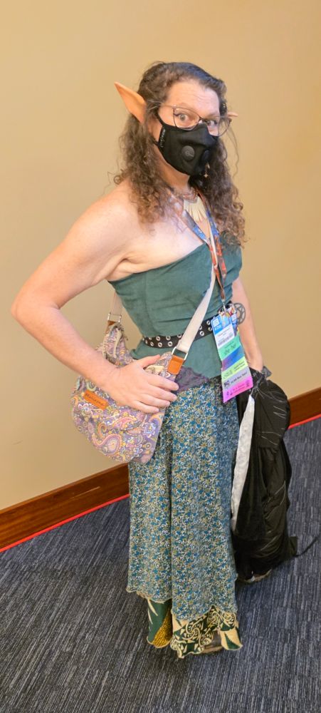 A masked woman woman with curly hair and long elf ears stands before a tan wall. She wears a green bodice, and green silk skirt. Her waist is belted and her shoulders are bare.