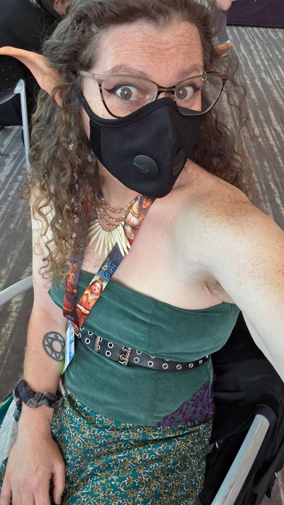A selfie of a masked woman with curly hair, glasses and elf ears. She wears a belted green bodice that leaves her freckled shoulders bare, patterned green silk skirt, conference lanyard, and necklace. She is seated with one hand resting on her lap and the other arm holding the camera.