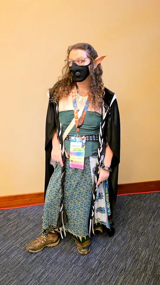 A curly haired woman in a green patterned skirt, green bodice, and black cloak wearing a black mask, GenCon badge, and elf ears looks down at the camera, one hand on her thigh, the other resting on a paisley crossbody bag.