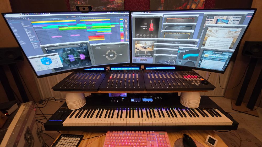 My studio workstation. At the top of the image are two large curved computer monitors, with an array of virtual instruments and digital audio workstation windows open on them. Underneath is a large midi controller that allows me to manage my mix with hardware instead of software. Beneath that is a full midi piano controller, and below that is a computer keyboard, mouse, and midi grid controller.