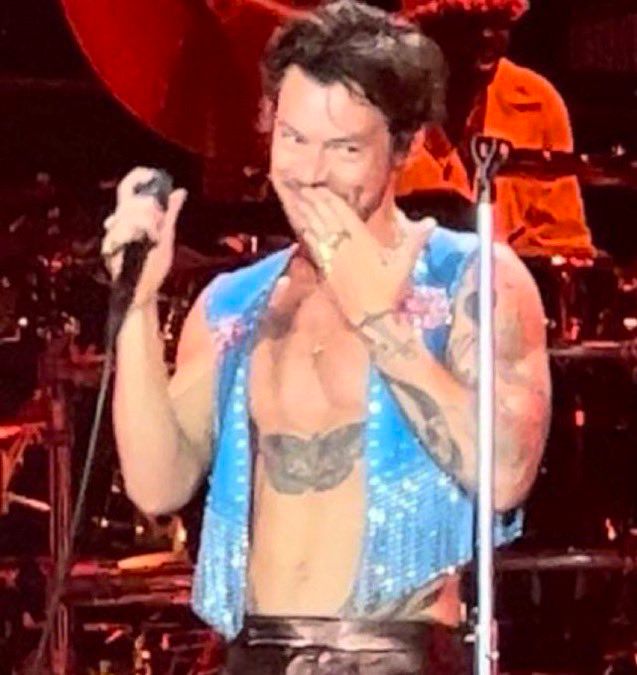 Harry Styles onstage in Barcelona with a mischievous expression. He wears glazed black leather trousers and a sparkly fringed GAY blue open vest with pink lollies on. 