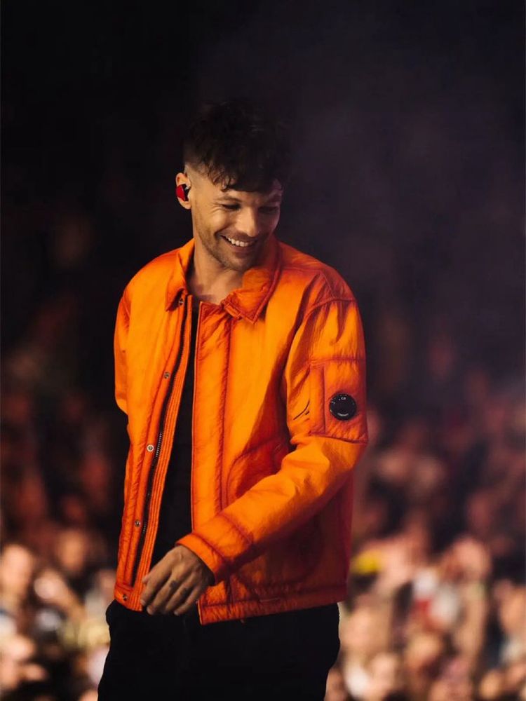 Beautiful Louis Tomlinson on stage at his music festival. He’s grinning and looking away from the camera. He wears black top and trousers with an orange jacket. 