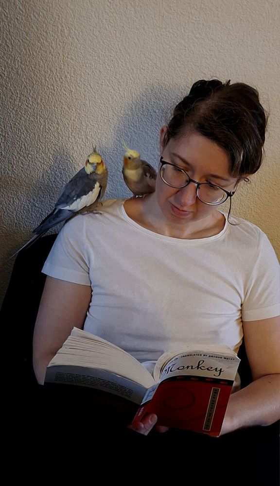 A woman reading a book with two cockatiels perched on her right shoulder