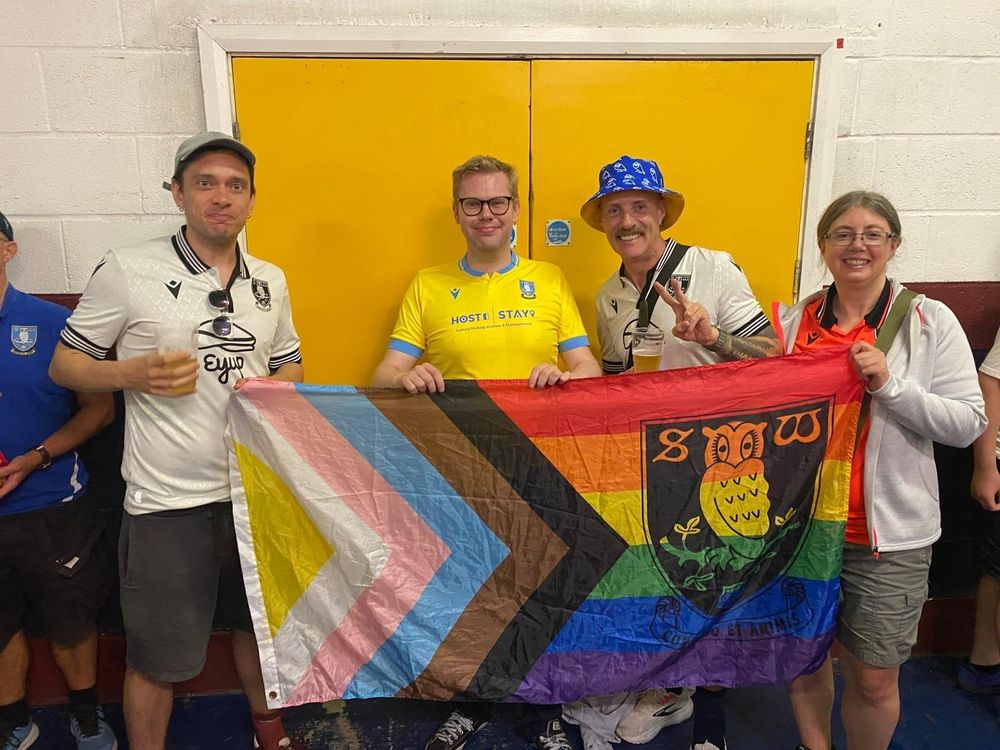 A photo of me, with three other Rainbow Owls members, holding Sheffield Wednesday's Progress Pride flag at Bolton Wanderers' stadium concourse.