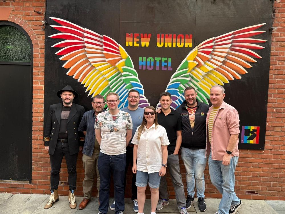 A photo of myself with Regional Alliance members from Rainbow Rams, Rainbow Spireites, Proud Stags, Rainbow Blades, Marching Out Together and Proud Forest standing together behind a New Union Hotel mural on Canal Street.