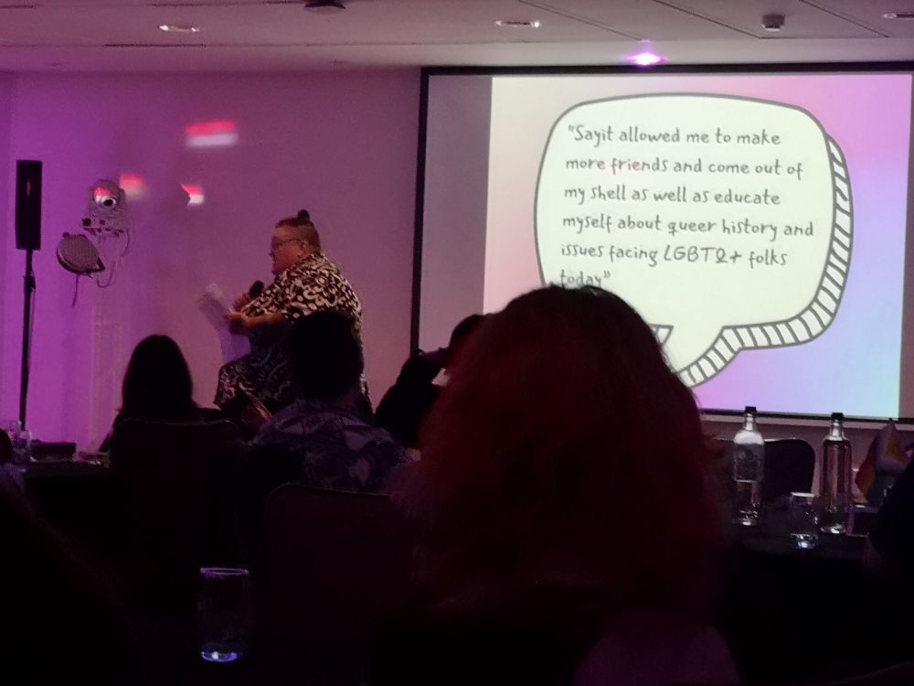 A photo of SAYiT's CEO Nicola Fearnley-Hill giving a speech at their 25th anniversary gala event. Nicola is placed behind a PowerPoint slide that states: "SAYiT allowed me to make more friends and come out of my shell, as well as educate myself about queer history and issues facing LGBTQ+ folks today."