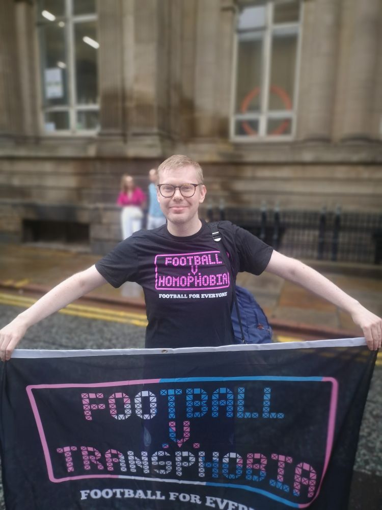 Photo of me holding the Football v Transphobia banner in a Football v Homophobia T-shirt at Leeds Pride.