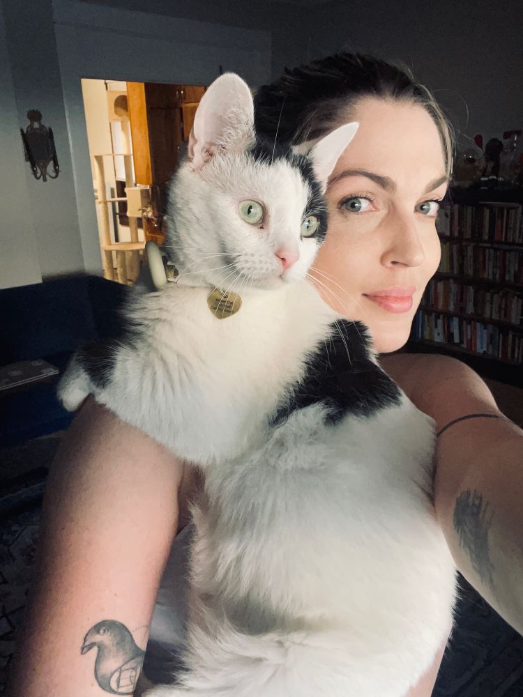 A white and black cat perched on a 30-something-year-old woman’s shoulder. The human is smiling and the cat looks alert and focused on something out of frame.