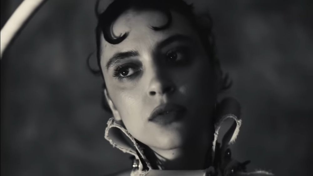 A picture of Pomme in black and white from her new single 'des excuses', with a pixie hairstyle and two hair piece curled at the front, looking to the side, off-camera. The collar of her outift is high, reaching her ears.