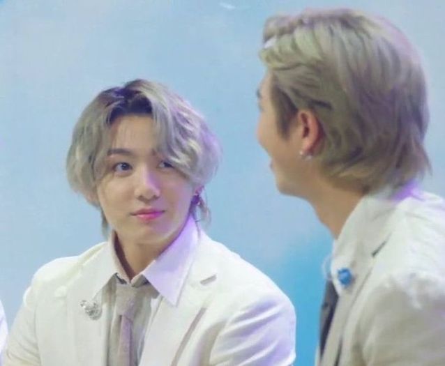 Jungkook with silver hair, a white suit and a tie, sitting on the left looking at Namjoon with adoring eyes. Namjoon is also wearing a white suit and has silver hair. He's looking past Jungkook with a smile.
