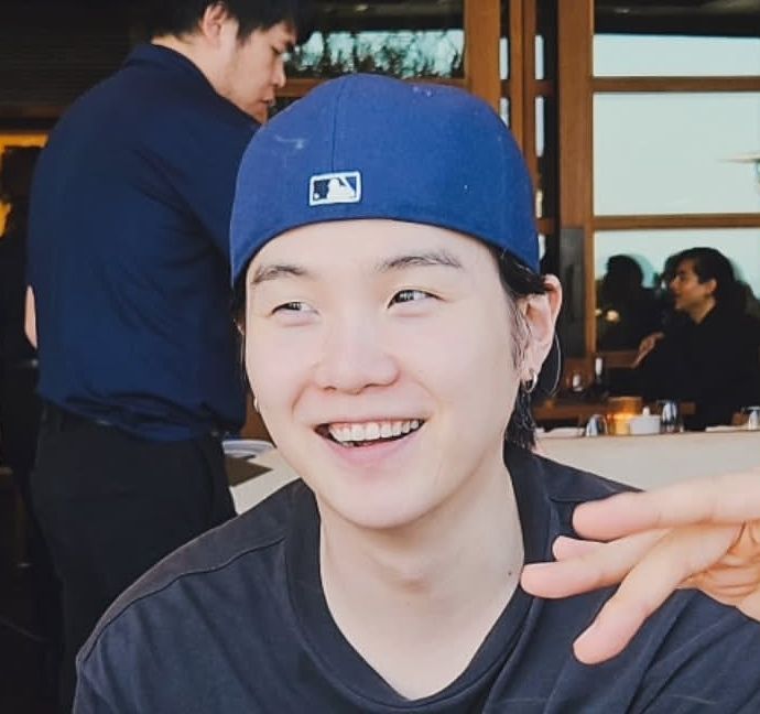 Yoongi smiling warmly like a dumpling, wearing a backwards blue baseball cap and a black t-shirt. He is indoors, with a blurred background of people and windows.