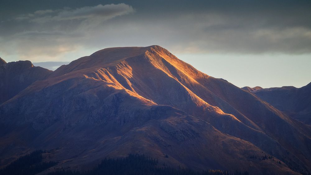 Soft sunrise light rakes across a rounded alpine peak, highlighting golden ridges and casting deep blue shadows on the surrounding mountains under a moody sky.