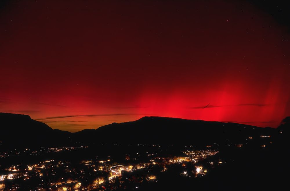Town lights sparkle in a dark mountain valley beneath an intense red aurora, its glowing bands rising above a thin stripe of green-yellow on the horizon and streaked with faint clouds.