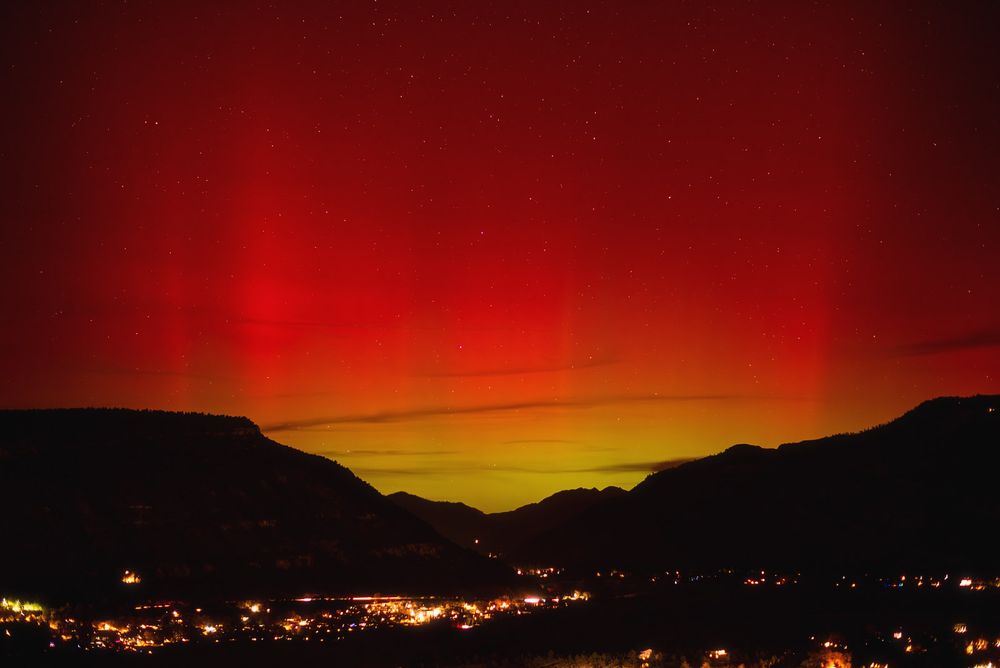 Nighttime mountain valley with a town’s lights below, under a vivid aurora: lime-green glow on the horizon fading into deep, ruby red curtains dotted with stars. Dark ridgelines silhouette the colorful sky.