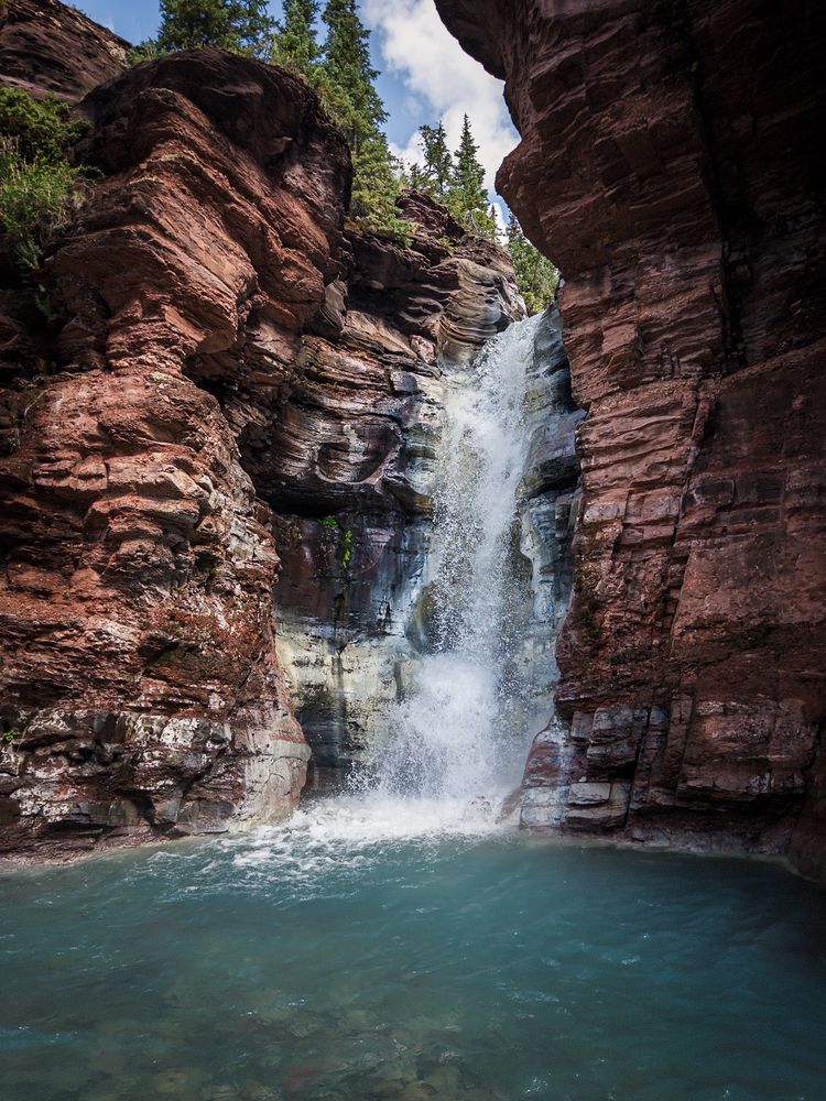 A waterfall cascades down layered red rock cliffs into a clear blue pool, surrounded by pine trees and under a partly cloudy sky.