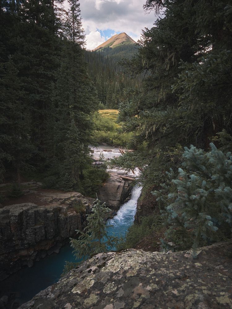A small waterfall with bright blue water flows through a rocky forested canyon, surrounded by dense evergreen trees, with a pointed mountain peak in the background under a partly cloudy sky.
