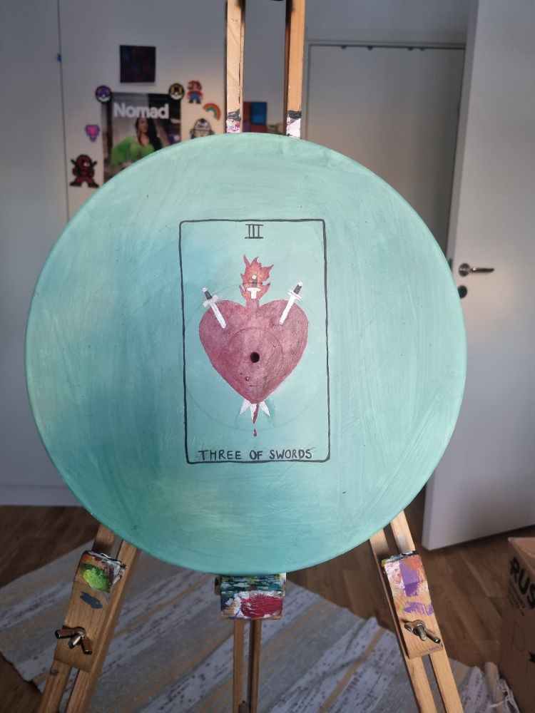 A painting on an old vinyl. There is a green/teal/mint background with a Tarot Card called "Three of Swords". There are three swords stabbing into the heart, and the heart bears resemblance to the sacred heart.