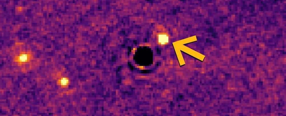 The newly discovered exoplanet is highlighted with an orange arrow.  The star it orbits is black in this image.  The remaining two bodies to the left are both background galaxies.