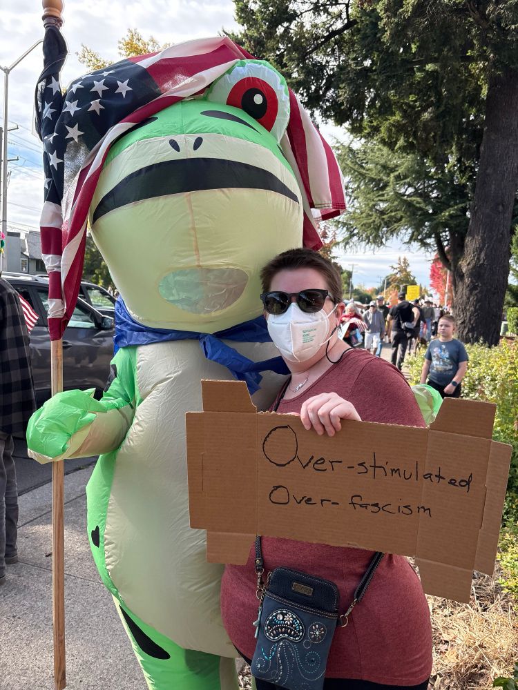 White woman in a mask. Her sign says Over-stimulated Over-fascism. She is standing with someone in an inflatable frog costume holding an American flag 