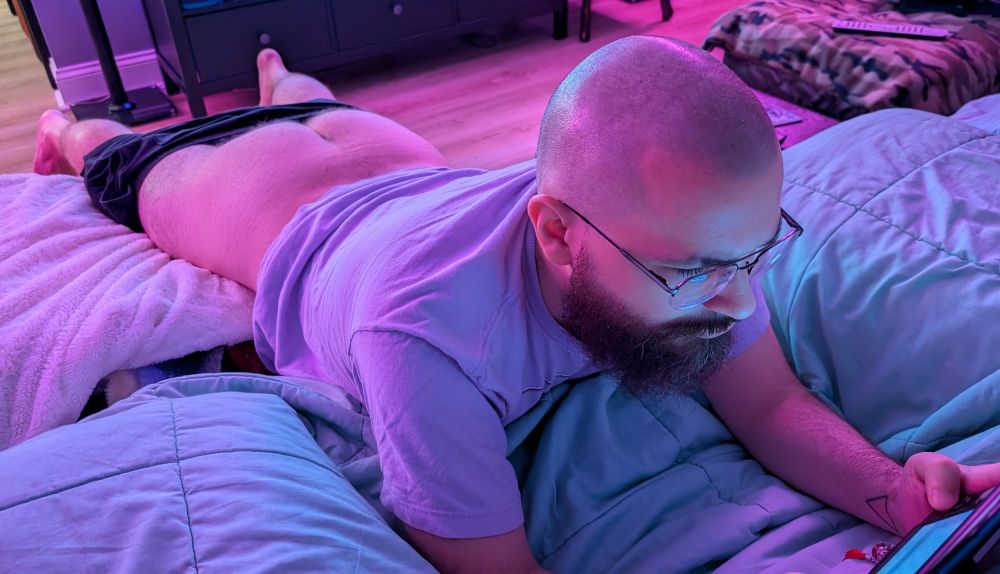 Hermit lays flat while playing SMT IV. His shirts are pulled down to show his bare ass. He is wearing a shirt. He is barefoot as always. 