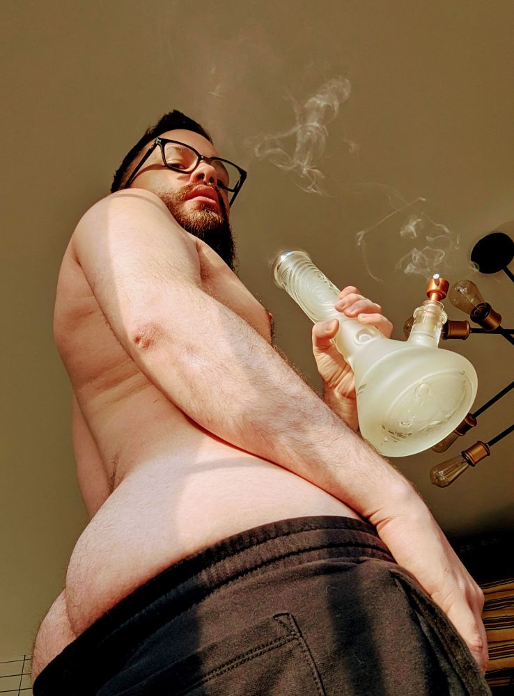 Adult Erotic Content: a shirtless man takes a hit of a bong. His shorts are pulled down to reveal the top of his ass cheeks. 