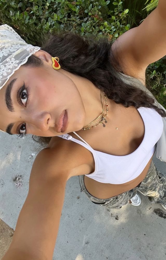Foto tirada de cima, Nour usa cropped de alça branco e uma bandana rendada branca, usa calça camuflada e está séria.