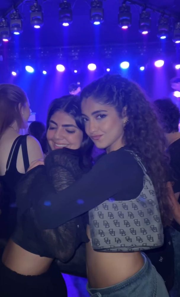 Nour está abraçando uma menina por trás, a menina usa calça preta e cropped preto de manga longa, sorrindo. Nour usa calça jeans e cropped preto de manga longa l, uma bolsa cinza no ombro, ela franze os lábios. Elas estão em um show.