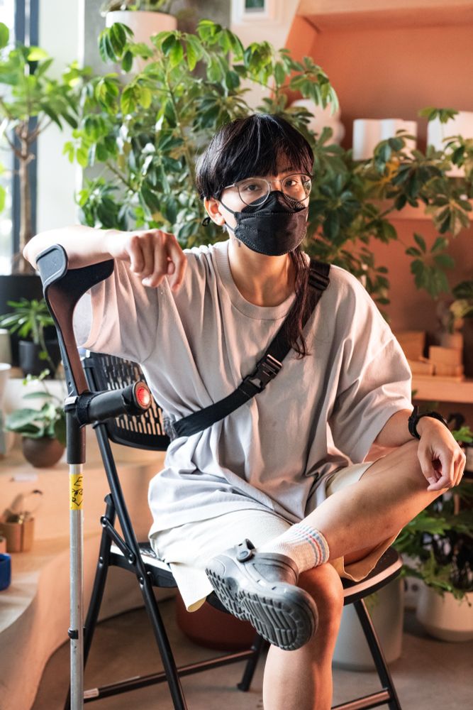 A masked non-binary Filipino person looks at the camera neutrally while sitting in a plant shop with his arm resting atop his forearm crutch. He has glasses, hair cut short and blunt save for a purposely wavy long strand, and is dressed in a large loose fit shirt, long shorts, and socks with clogs.