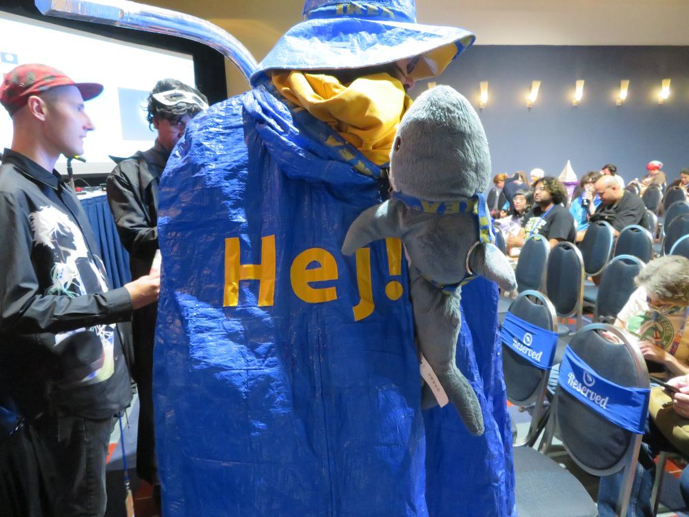 Ikea Wizard cosplayer whose cape says "Hej!" and has a small Blahaj on his back.