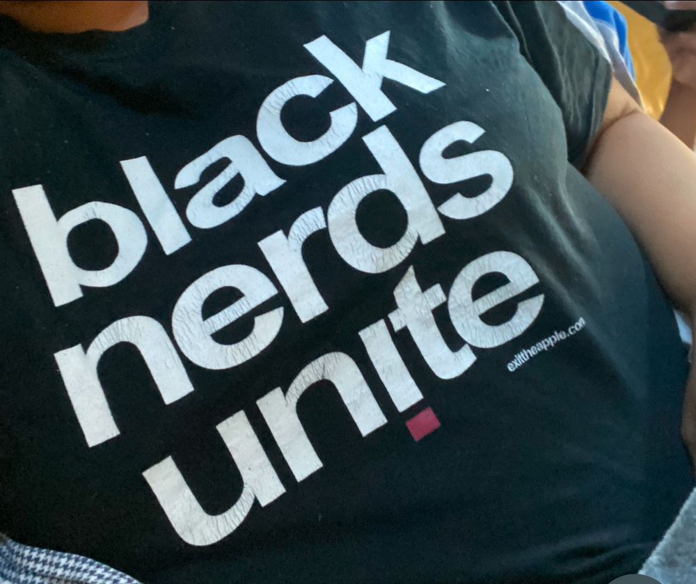 Picture of me in my car wearing a black tshirt with white writing in English that says, “Black Nerds Unite,” a gift from a former student ♥️