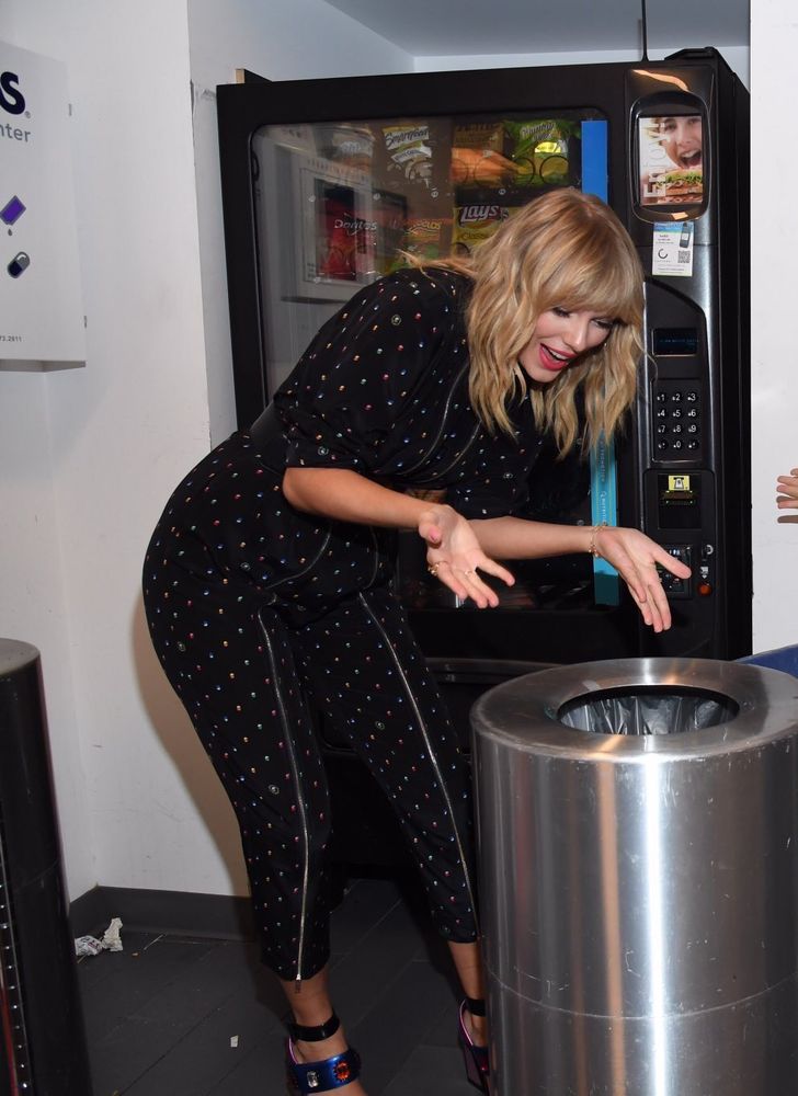Taylor swift with short blonde hair in a black jumpsuit with colorful dots all over it leaning over a trash can, holding her hands out over it and smiling. A vending machine is behind her