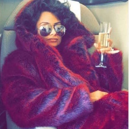 A Black woman sits in a plush seat in a plusher magenta fur coat with shades on. She elegantly holds a glass of champagne. 