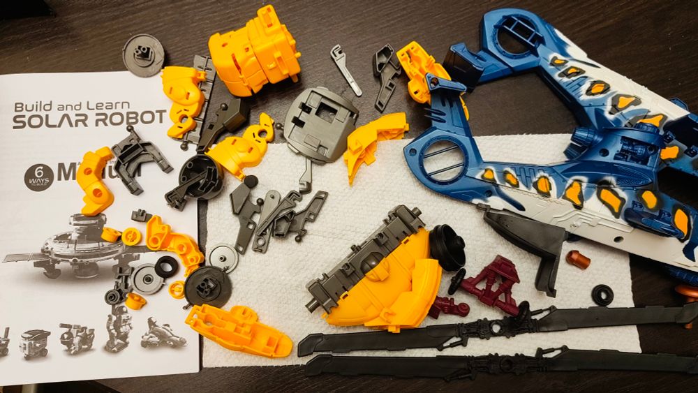 Yellow and gray plastic parts are spread out on a table next to their manual for a "Build and Learn Solar Robot" kit. Beside it is a disassembled toy helicopter, mostly blue and white.