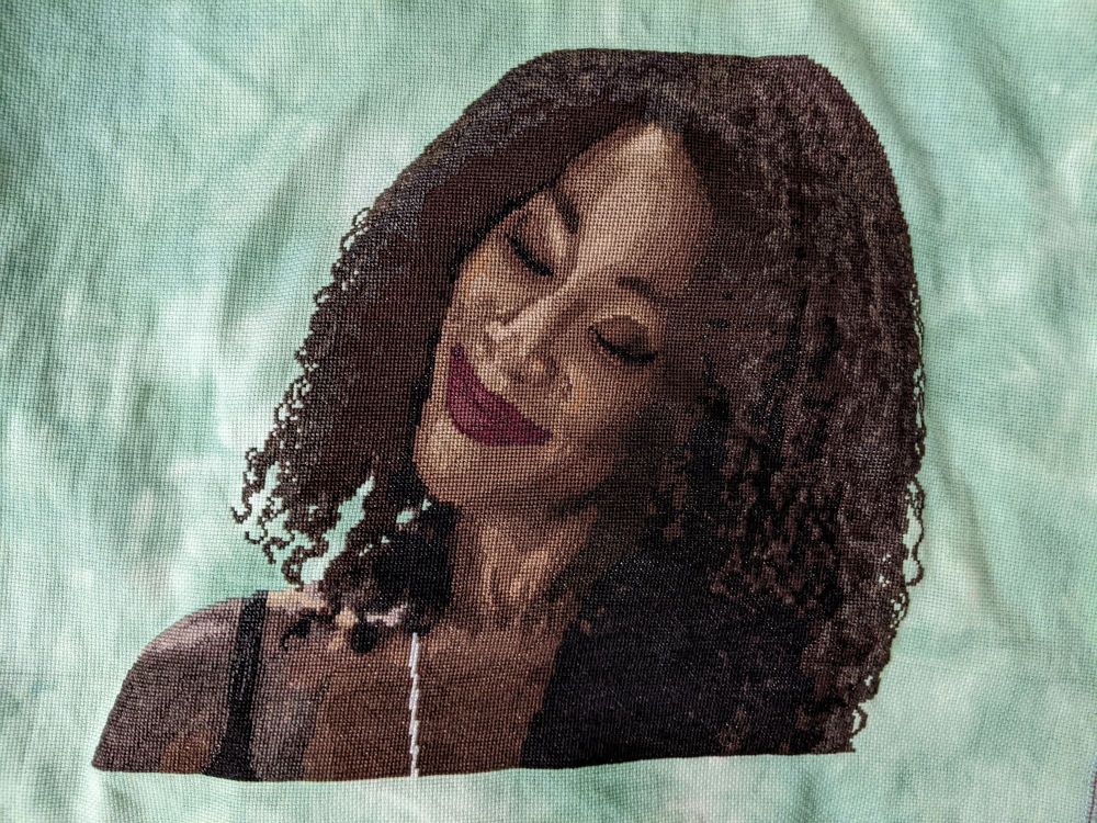 A photorealistic cross stitch of Death from the Sandman TV show. It's a black woman shown from the shoulders up. Her head is tilted to one side and her eyes are closed as she smiles warmly. The background is a mottled green hand-dyed fabric.