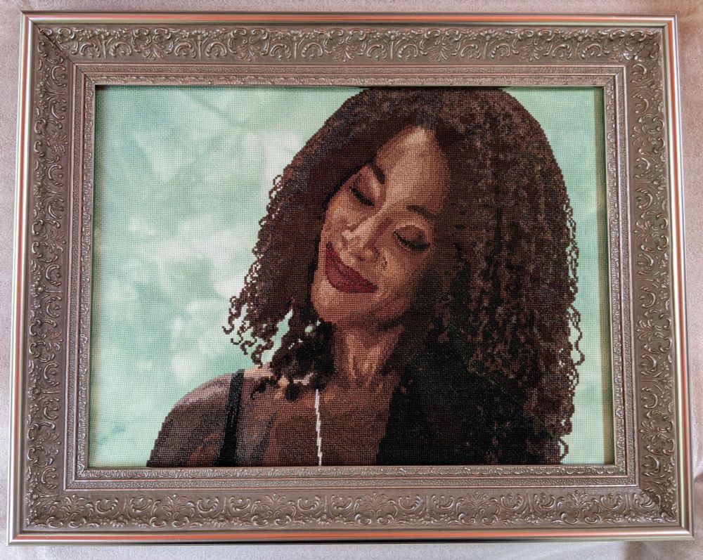 A cross stitch piece of Death from The Sandman. She's a black woman with curly, shoulder-length black hair, smiling warmly with her eyes closed and her head tilted to one side. She's done in a photorealistic style on mottled green Aida fabric. The frame is thick and silver with a filigree-style pattern on it.