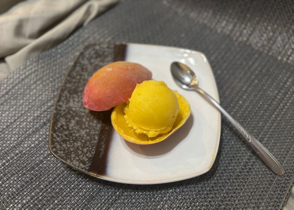A square plate on a grayish placemat presenting a scoop of mango sorbet in a frozen, hollowed-out mango shell with a long dessert spoon.