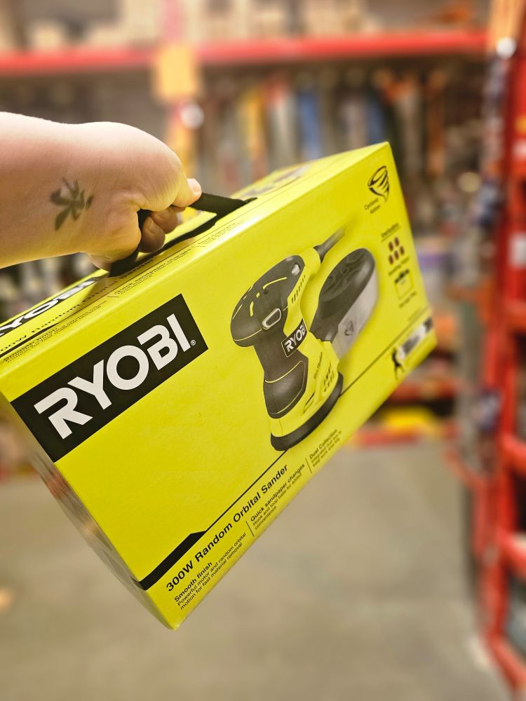 A hand holds a yellow box containing a Ryobi orbital sander.