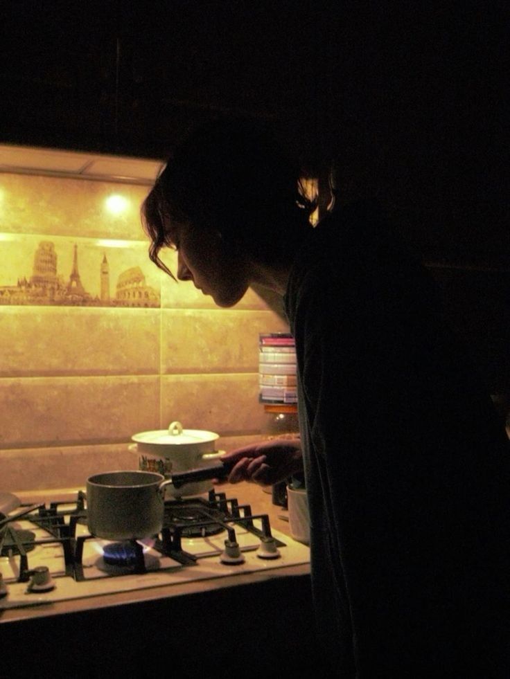 profile of a person is backlit by the light above stove. they're holding a small pot