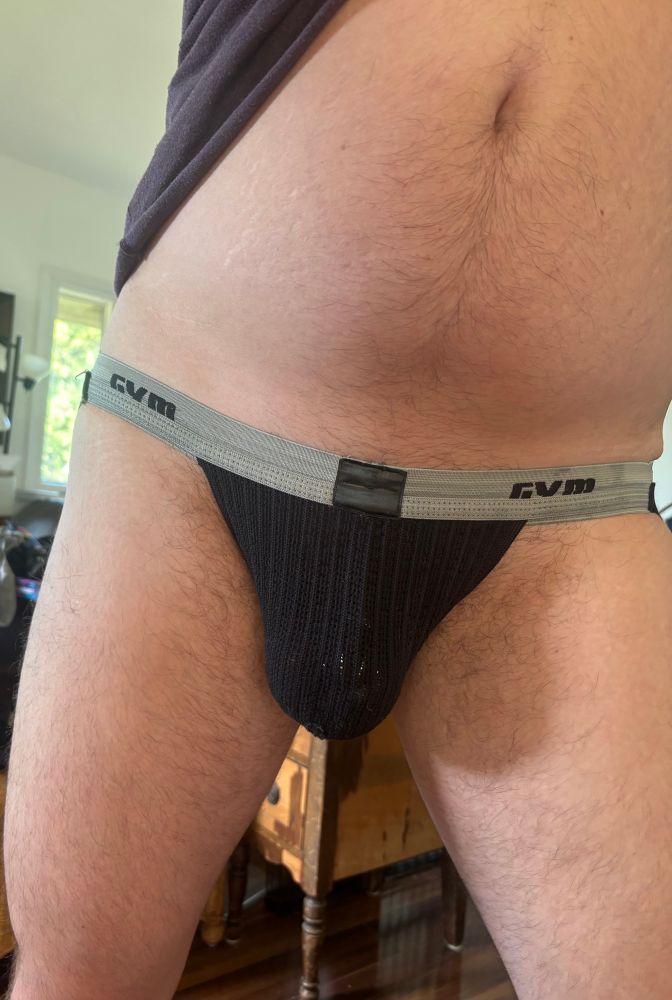 a close-up photo of a fair skinned man’s lower tummy, crotch and thighs. he’s wearing a black GYM brand jock strap with a grey waistband and the silver bars of a chastity cage are visible through the semi-transparent mesh pouch. 